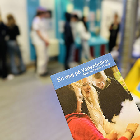 Broschyr med titel "En dag på Vattenhallen" i fokus, i bakgrunden skymtas årskurs 8 elever på Vattenhallen. Foto. 